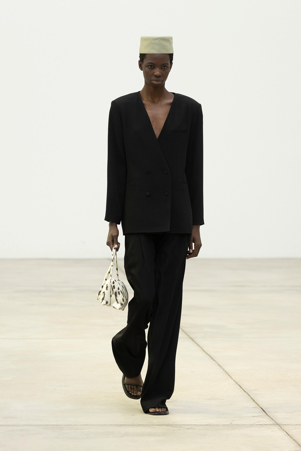 Model walking a runway in a black double-breasted blazer with matching wide-leg pants, a pale hat, and a small white patterned handbag.