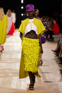Model walks the runway in a bright yellow cape-style top with a white center and a textured yellow feather skirt, complemented by a purple headwrap and a small beige handbag.
