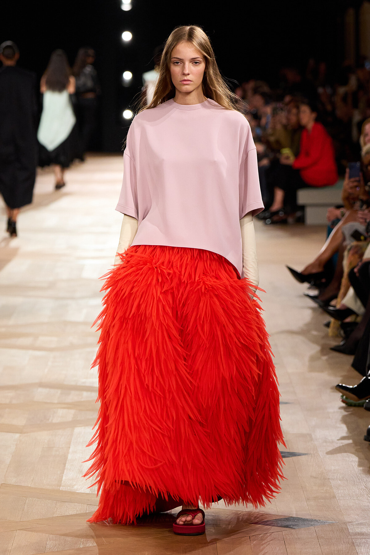 Model walks the runway in a soft pink oversized top and a voluminous red feathered skirt, audience in the background.