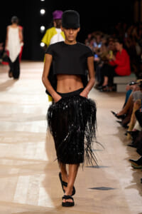 Model walking the runway in a black cropped top and long black fringe skirt, paired with a tall black hat and sandals.