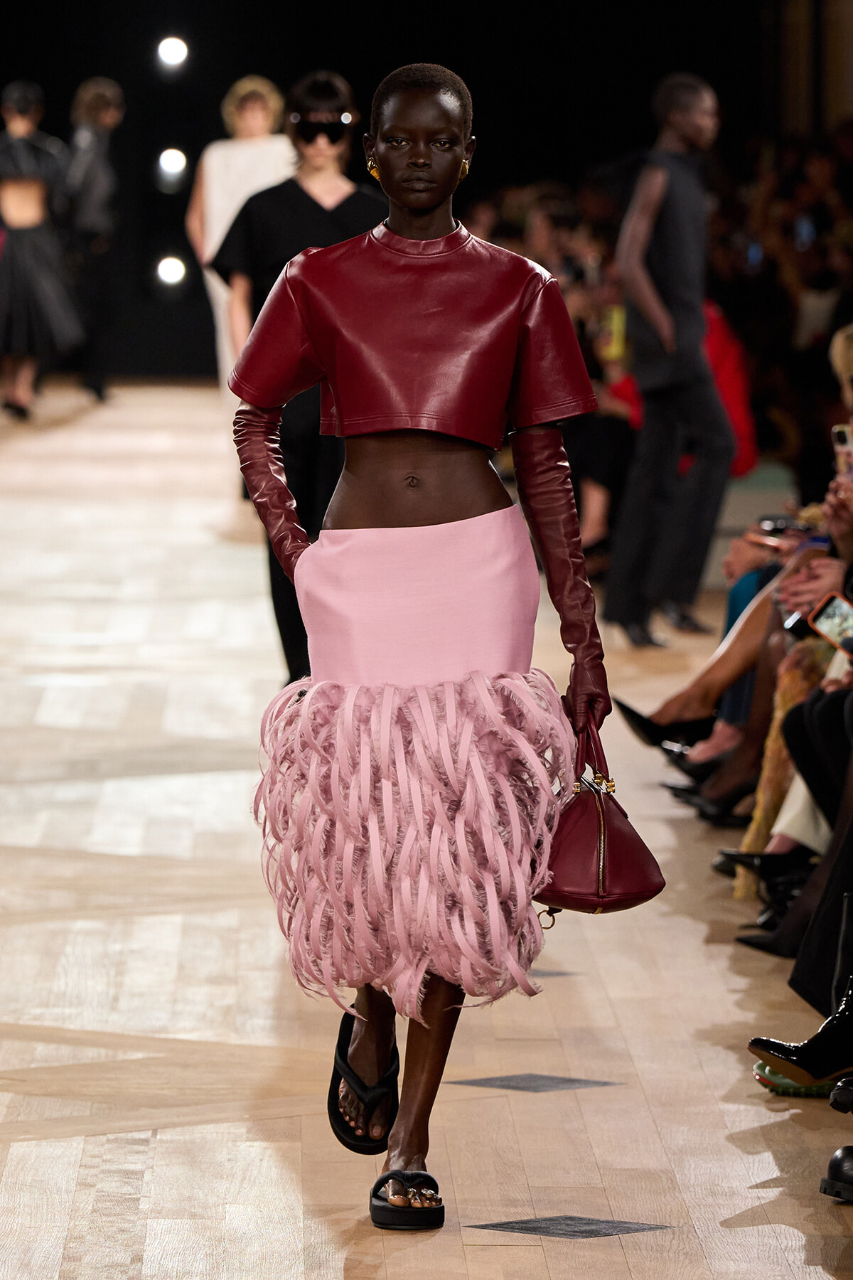 Model walks the runway in a burgundy cropped leather top with matching long gloves and a pink fringed skirt, carrying a burgundy handbag.