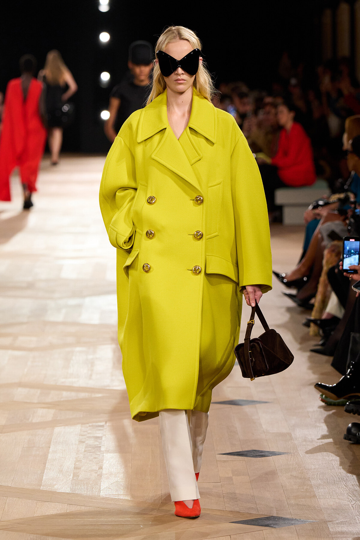 Model wearing a bright lime oversized double-breasted coat with wide lapels, white trousers, orange shoes, and large black sunglasses, carrying a small brown handbag on a fashion runway.