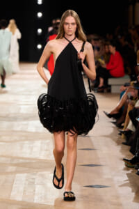 Model walking the runway in a black halter-neck dress with a voluminous fringe hem, carrying a shoulder bag.