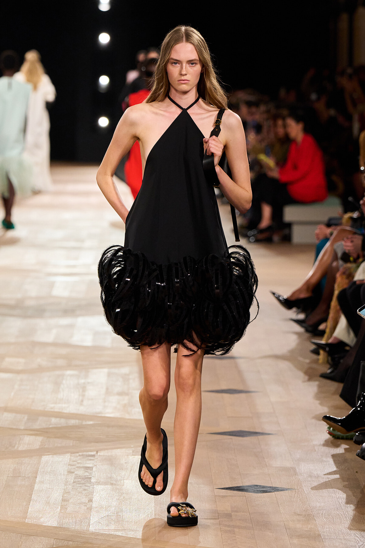 Model walking the runway in a black halter-neck dress with a voluminous fringe hem, carrying a shoulder bag.