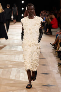 Model walks a fashion runway wearing a knee‑length ivory dress covered in 3D floral appliqués, with black gloves and flat sandals on a wooden catwalk.