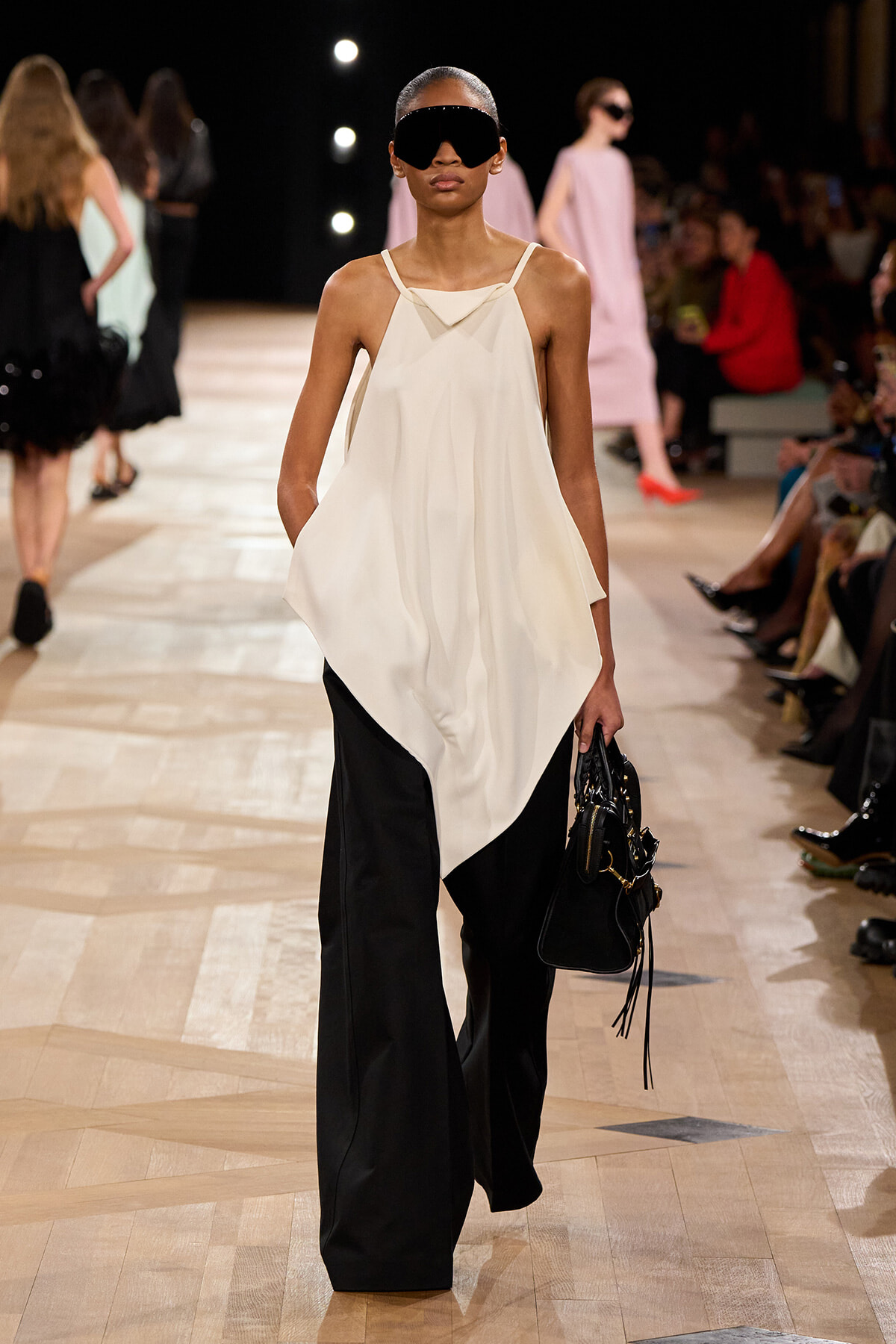 Model on a fashion runway wearing a white draped top over black wide-leg pants, oversized black sunglasses, and a black handbag.