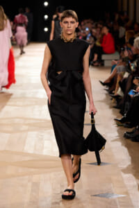 Model walks the runway in a black sleeveless midi dress with a bow at the waist and a chunky gold necklace.