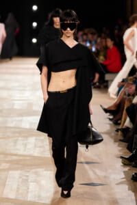 Model walks the runway in a black cropped top with cape overlay and matching black trousers, holding a black handbag with gold hardware at her side.