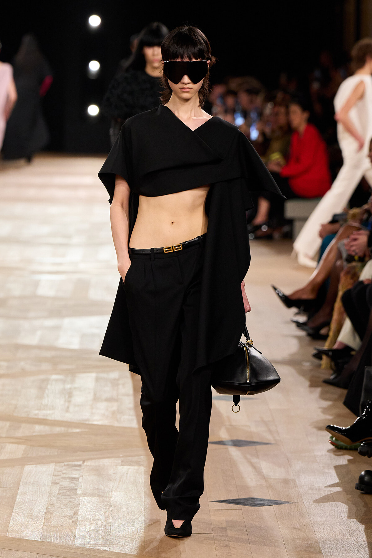 Model walks the runway in a black cropped top with cape overlay and matching black trousers, holding a black handbag with gold hardware at her side.
