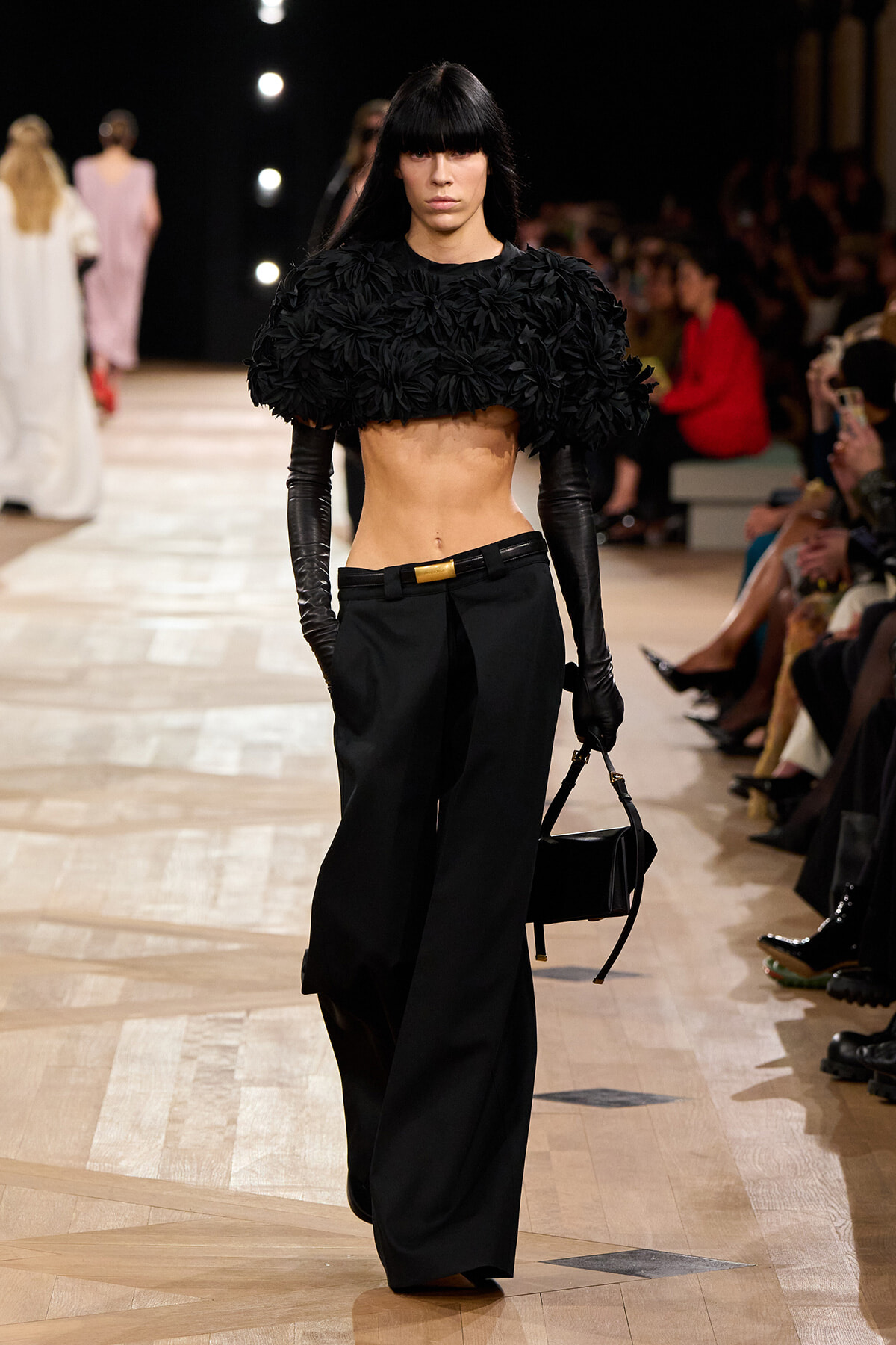 Model on runway wearing a black feathered crop top, long black gloves, wide-leg black trousers, and a small black handbag.