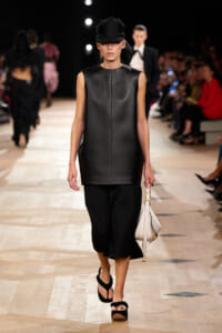 Model on a fashion runway wearing a black sleeveless leather tunic, black cap, cropped wide-leg pants, black platform sandals, and a white handbag