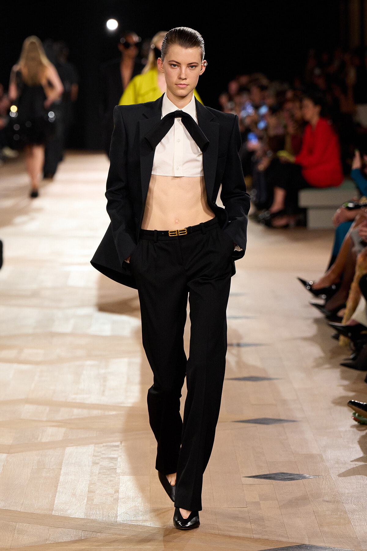 Model on a runway wearing a black tailored suit with a white cropped shirt and a large bow tie, hands in pockets.