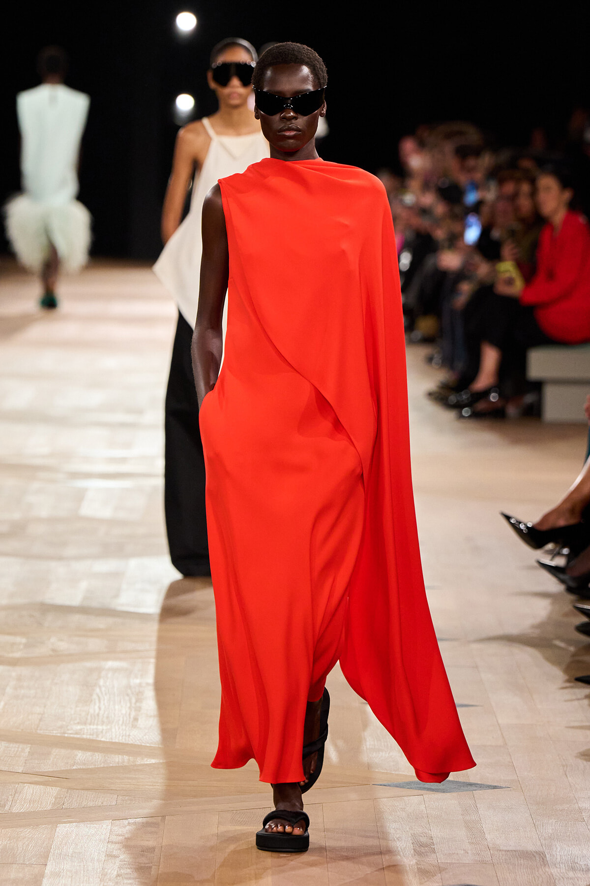 Model walks a runway wearing a bright red, one-shoulder cape dress with black sunglasses and sandals.