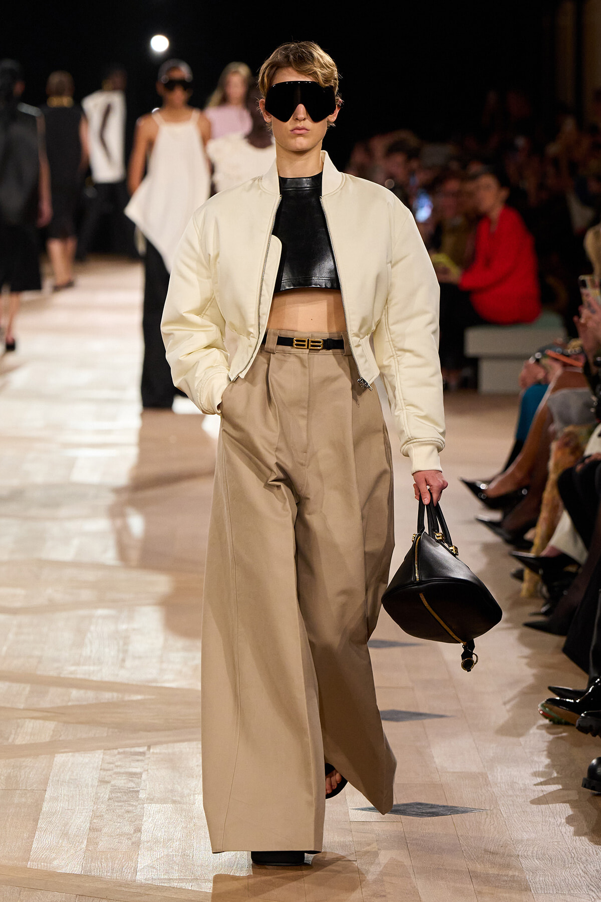 Model on a runway wearing a cream bomber jacket over a black crop top, tan wide-leg trousers, oversized sunglasses, and a black structured handbag.