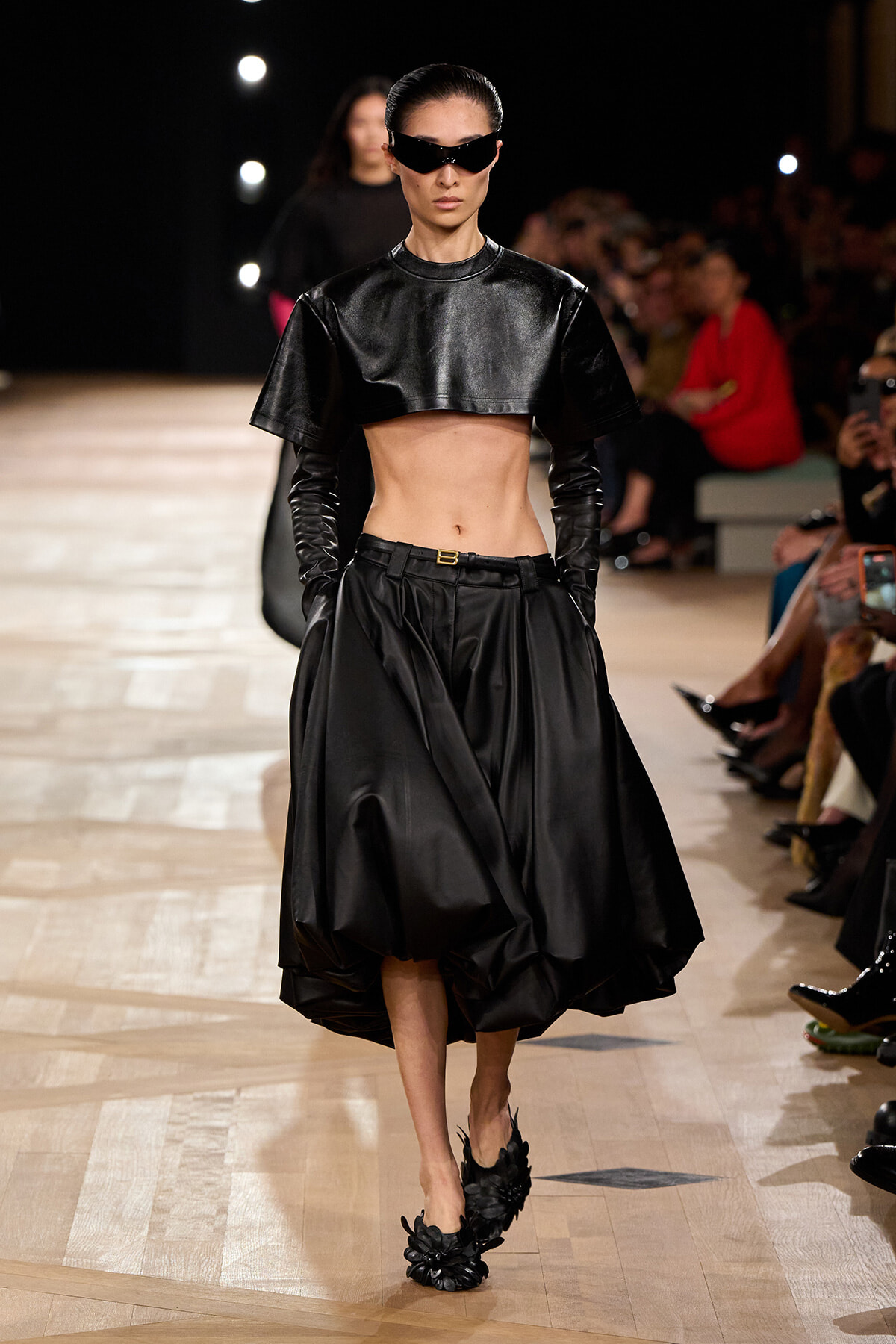 Model walks a runway in a black cropped leather top, voluminous midi skirt, long gloves, and oversized sunglasses; floral-embellished heels complete the look.