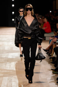 Model walks the runway in an oversized black leather wrap top, black trousers, and oversized black sunglasses, carrying a small black handbag.