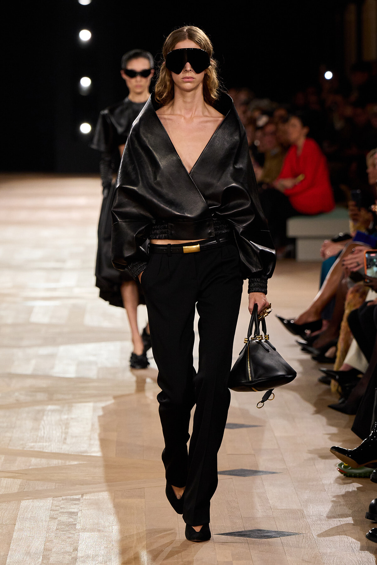 Model walks the runway in an oversized black leather wrap top, black trousers, and oversized black sunglasses, carrying a small black handbag.