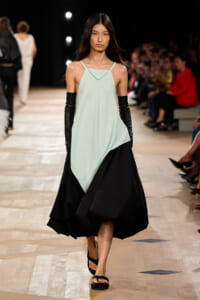 Female model walks a fashion-show runway wearing a mint green sleeveless top with an asymmetric black skirt, long black gloves, and black sandals.
