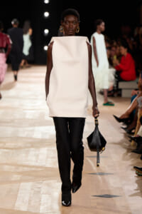 Model walking a runway in a minimalist ivory sleeveless tunic with black pants, gold hoop earrings, and a small black handbag