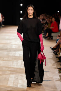 Model walking a fashion runway in an oversized black outfit with bright pink gloves and a hot-pink handbag.Colleague audience blurred behind her as she strides down the catwalk seeks focus on accessories.