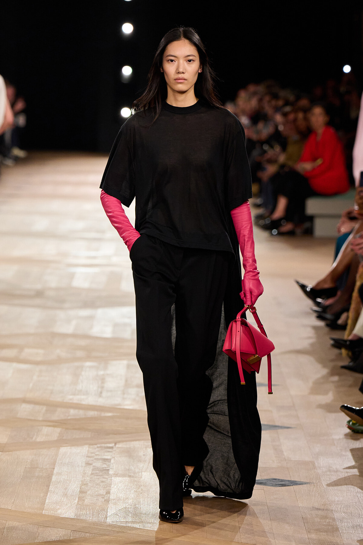 Model walking a fashion runway in an oversized black outfit with bright pink gloves and a hot-pink handbag.Colleague audience blurred behind her as she strides down the catwalk seeks focus on accessories.