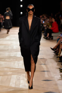 Model walking a runway in a long black blazer dress with a deep V, no shirt, oversized shoulders, and a thigh-high slit, paired with glossy black heels and large black sunglasses.