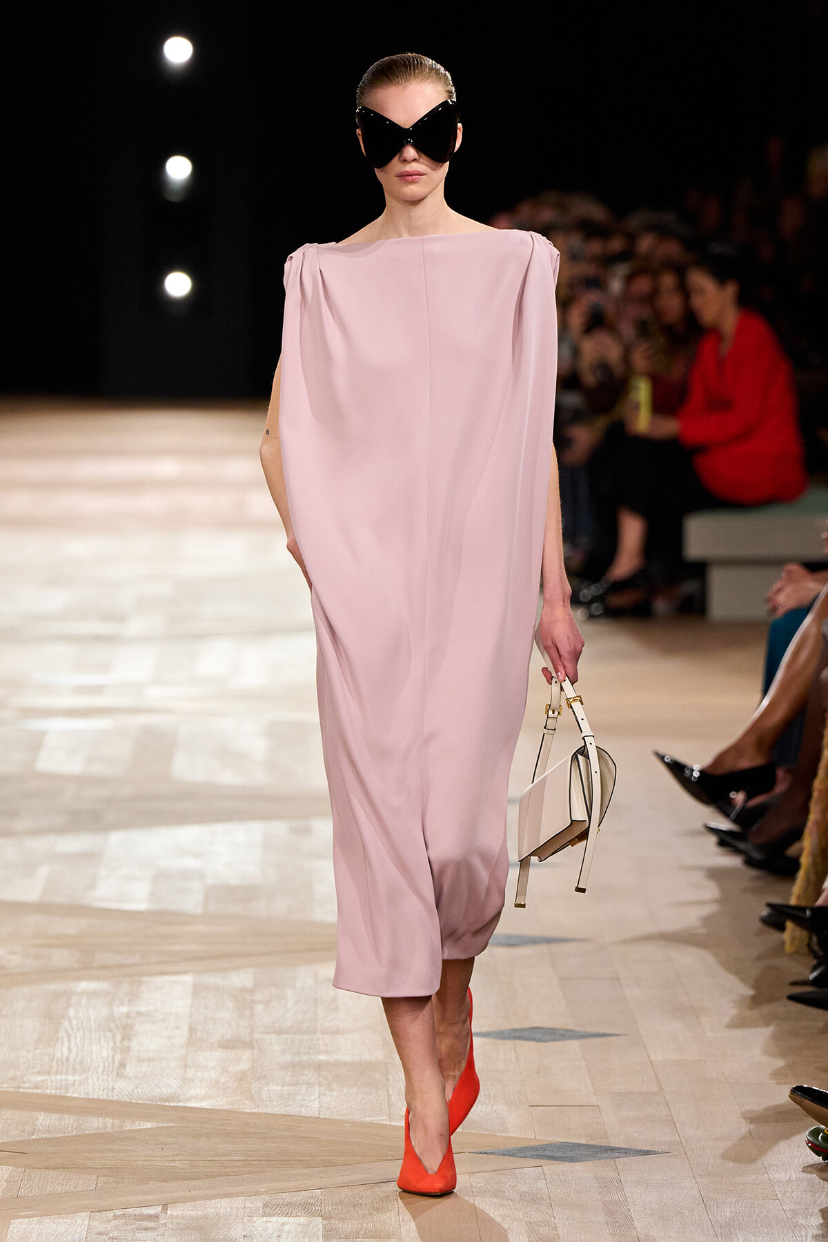 Model walking the runway in a pale pink draped dress, oversized black sunglasses, bright orange heels, and a small white handbag.
