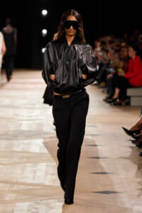 Model walking a runway in a glossy black oversized leather jacket with large sculpted sleeves and hands in pockets, paired with black tailored pants and a slim belt, plus oversized dark sunglasses.