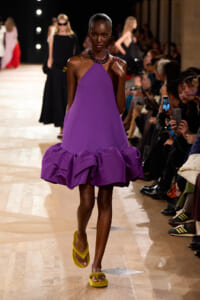 Model walks the runway in a sleeveless purple halter dress with a voluminous ruffled hem, paired with bright yellow fuzzy sandals.