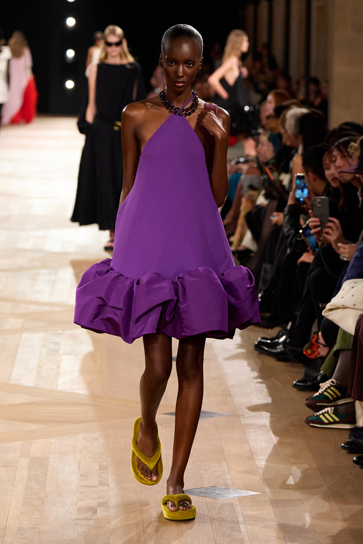 Model walks the runway in a sleeveless purple halter dress with a voluminous ruffled hem, paired with bright yellow fuzzy sandals.