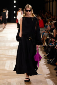 Model walking a runway in a black maxi dress with a cape-like, one-shoulder sleeve, cinched at the waist with a belt, carrying a bright pink handbag; wearing sunglasses and black sandals, with seated audience along the catwalk.