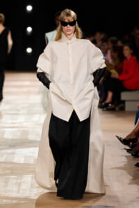 Model walks the runway in an oversized white button-down shirt over wide black trousers, paired with black gloves and angular sunglasses.