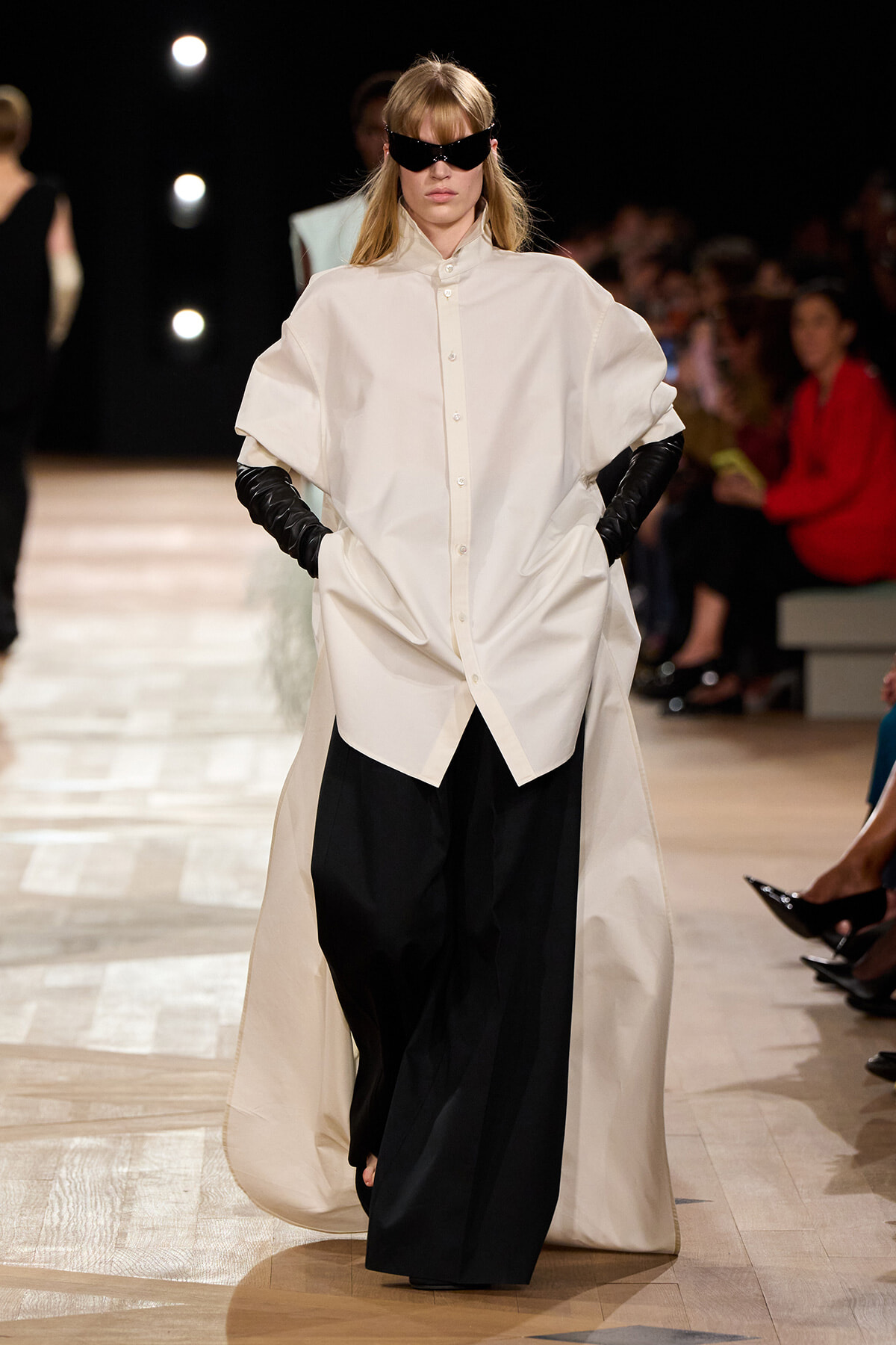 Model walks the runway in an oversized white button-down shirt over wide black trousers, paired with black gloves and angular sunglasses.