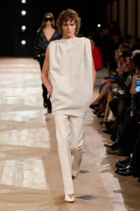 Model walks the runway in a minimalist cream sleeveless top and matching straight pants at a fashion show.