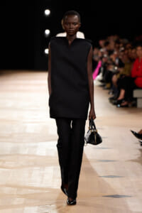 Model walking down a runway in an all-black ensemble—a sleeveless tunic and tailored trousers, carrying a small black handbag.