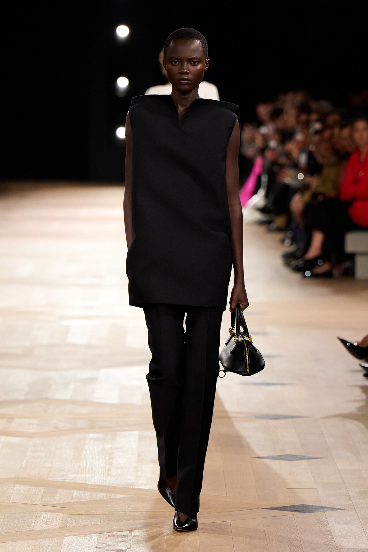 Model walking down a runway in an all-black ensemble—a sleeveless tunic and tailored trousers, carrying a small black handbag.