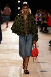 Model on a fashion runway wearing a dark olive-green leafy jacket, a blue denim skirt, black sandals, and a red handbag.