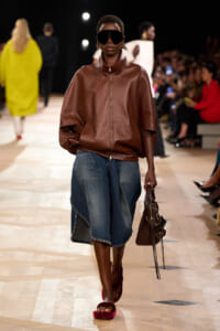 Model on a runway in an oversized brown leather jacket, knee-length denim skirt, giant black sunglasses, red sandals, and a brown handbag.