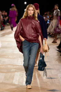 Model walks a fashion runway wearing a burgundy leather cape jacket with high collar and blue jeans, carrying a blue handbag.
