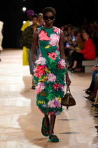 Model walks a runway in a bright, floral feathered dress with green and pink hues, dark sunglasses, green sandals, and a small brown handbag.