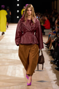 Model walks the runway in a burgundy leather belted jacket, brown wide-leg pants, and bright pink heels, carrying a brown bag.
