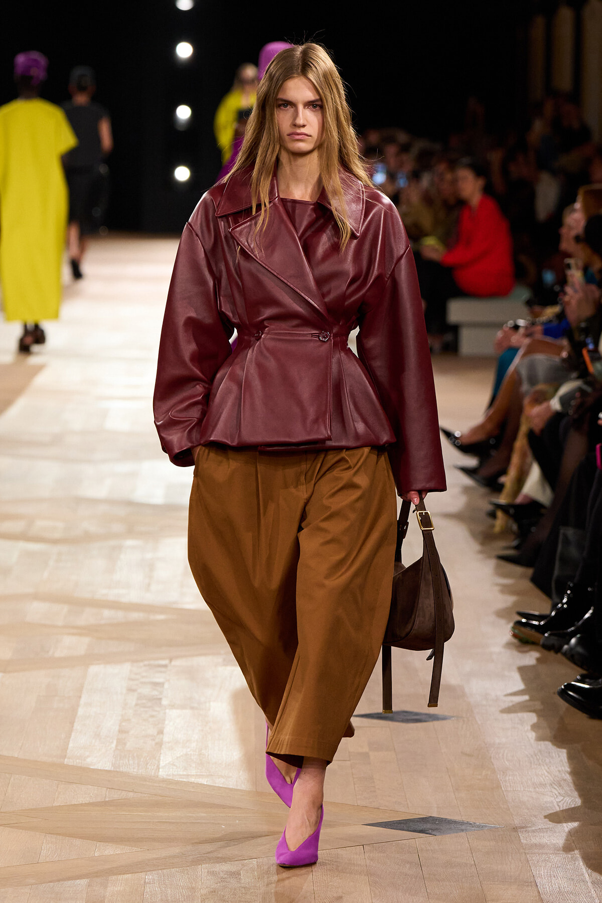 Model walks the runway in a burgundy leather belted jacket, brown wide-leg pants, and bright pink heels, carrying a brown bag.
