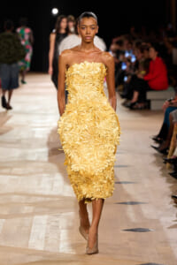 Model walking the runway in a strapless yellow dress covered in large flower appliqués; beige pumps and a focused expression, audience in the background.