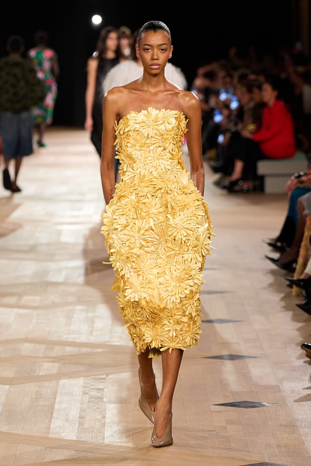 Model walking the runway in a strapless yellow dress covered in large flower appliqués; beige pumps and a focused expression, audience in the background.