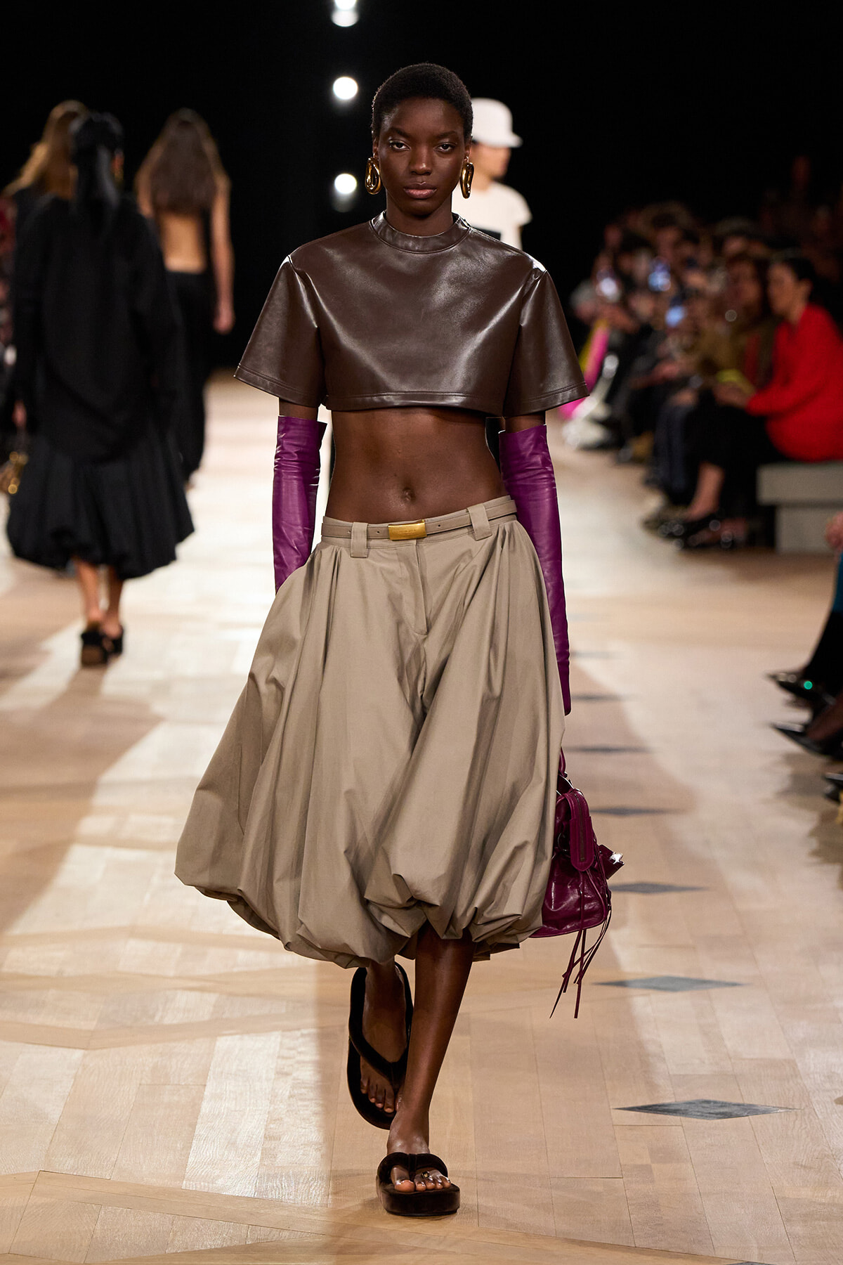 Model walks the runway in a brown leather crop top, oversized beige balloon skirt, purple gloves, and brown sandals while carrying a purple bag.