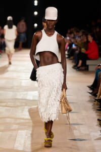 Model walks a runway in a white cropped top, feathered midi skirt, white cap, and tan handbag; yellow platform sandals visible