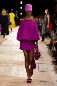 Model on a runway wearing a bold magenta off-the-shoulder top with a matching mini skirt, tall magenta hat, sandals, and a purple handbag.