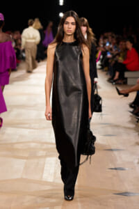 Model walking the runway in a sleeveless black leather dress with an asymmetric hem, carrying a black handbag.