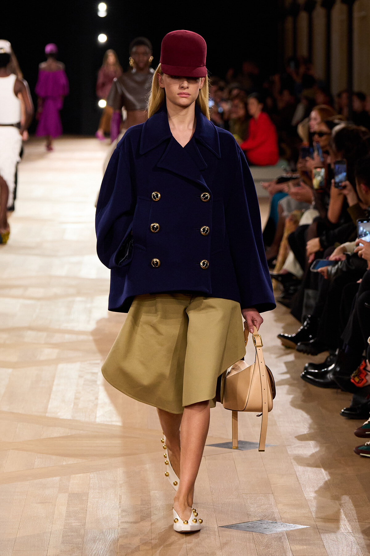 Runway model wearing a navy double-breasted coat with gold buttons, a tan knee-length skirt, a burgundy hat, white studded flats, and carrying a beige handbag; audience along the runway in the background.