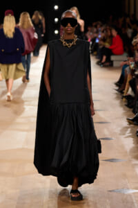 Model on runway wearing black sleeveless cape top and voluminous skirt, oversized sunglasses, chunky gold necklace, and sandals.